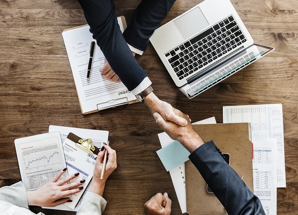 Poignée de main entre deux professionnels en costume autour d’une table de réunion avec ordinateur portable, documents et graphiques financiers.