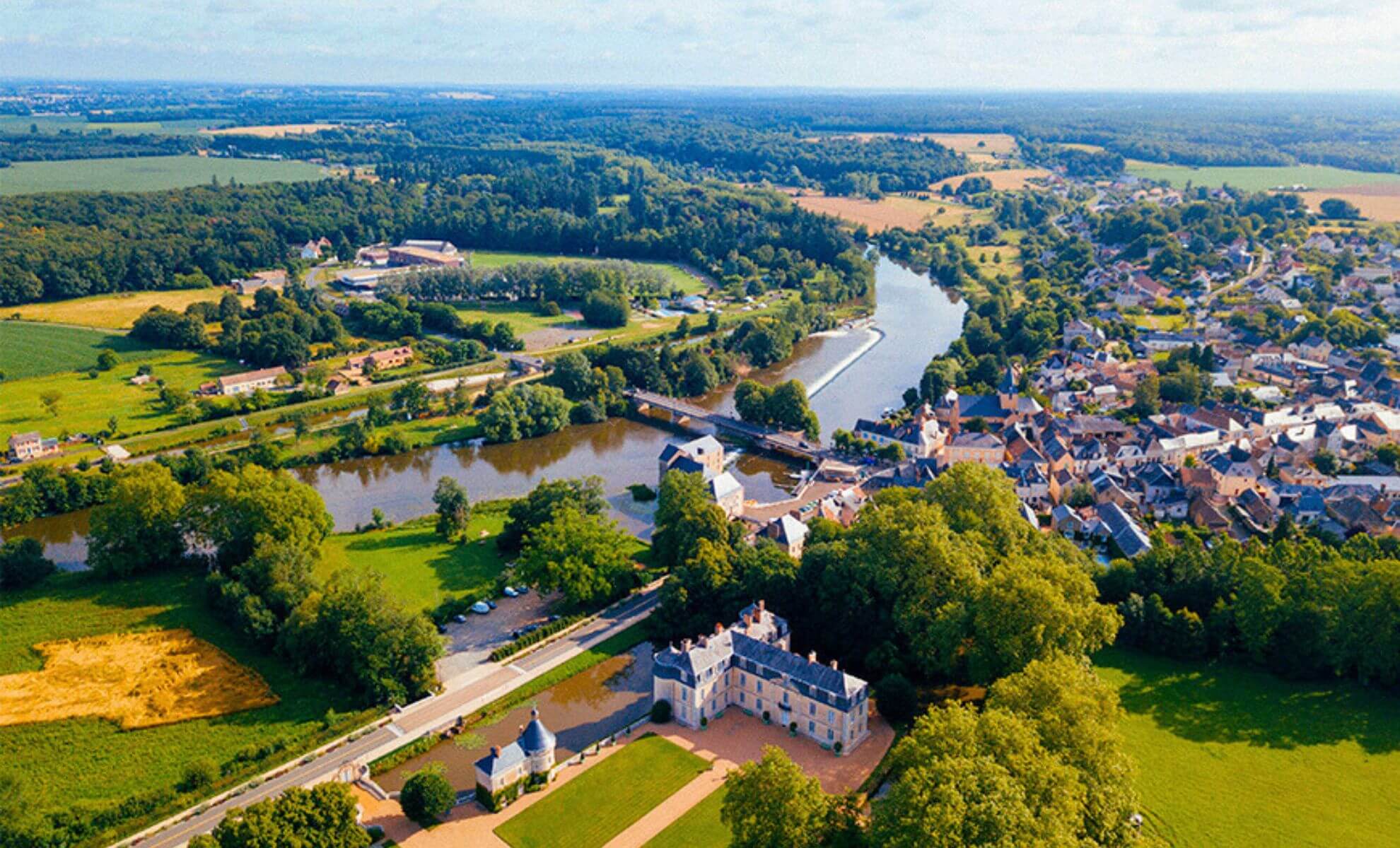 Vue aérienne d’un village dans la vallée de la Sarthe, avec une rivière sinueuse, un château, des champs et une campagne verdoyante.