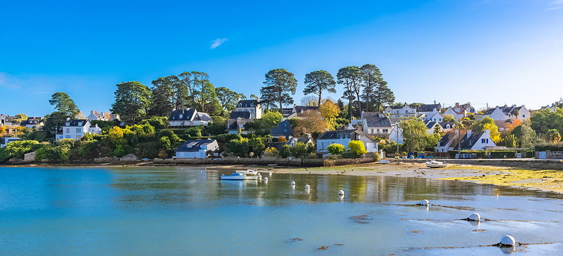 Village côtier dans le Morbihan, avec des maisons traditionnelles entourées de verdure et une mer calme au premier plan.